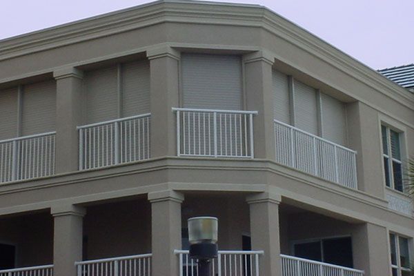 Tan building with balconies, white railings, and closed beige roll-down shutters.