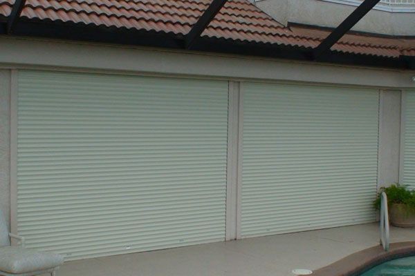 Closed beige roll-up shutters on an outdoor patio, under a brown-tiled roof.