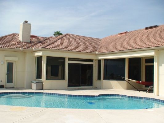Poolside view of a house with a tiled roof, pool, and a hammock. Beige exterior, windows, and clear sky.