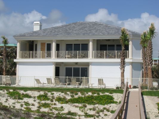 Two-story white beach house with columns, balcony, and gray roof, sitting on sand dunes with palm trees.