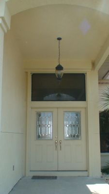 Double cream-colored entry doors with glass panels and a transom window under a covered porch. A hanging light is above.