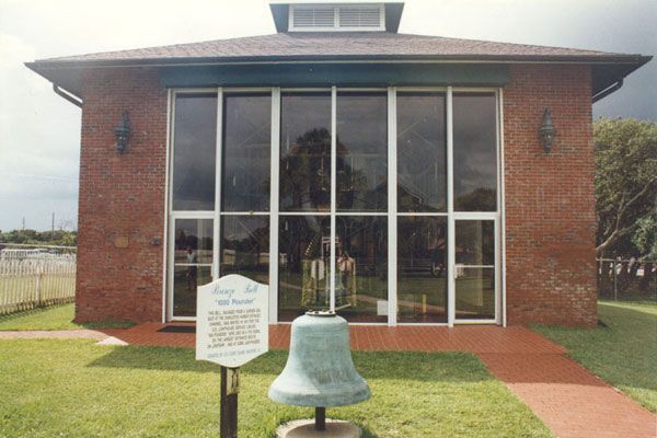 Brick building with large windows; a bell sits on the lawn. A sign is near the bell.