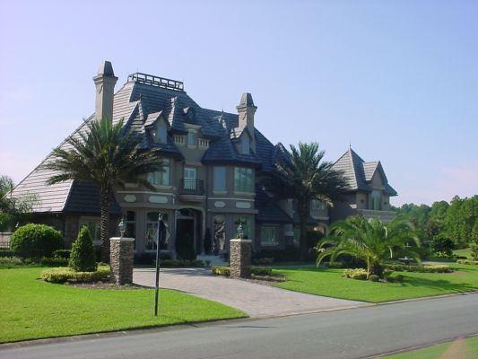 Large gray mansion with a slate roof, palm trees, and green lawn, in an upscale suburban setting.