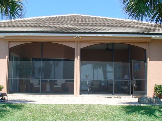 Screened-in patio with arched openings, beige walls, and brown roof. Green grass in foreground.