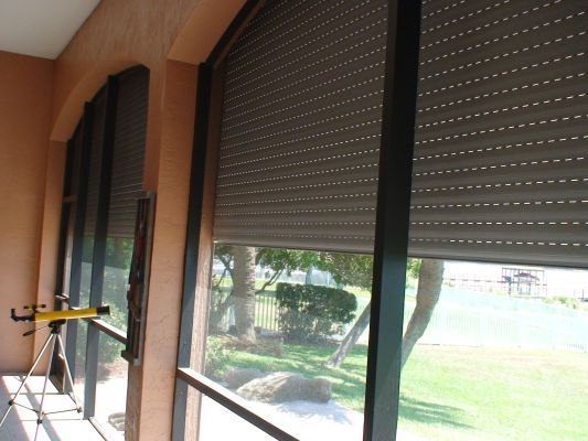 Brown roller shutters partially covering a window, with a view of greenery outside.