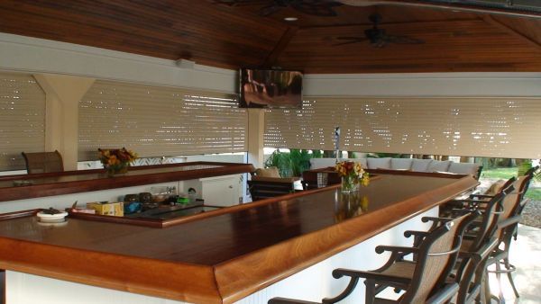 Outdoor bar with wooden counter, bar stools, and TV, under a covered patio.