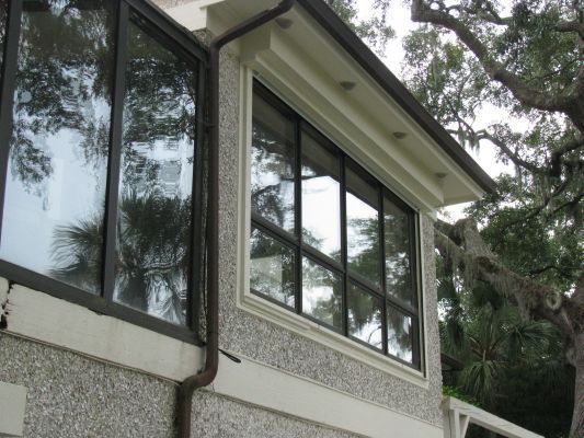 Exterior house corner with windows, light stucco, and dark trim. Tree branches with Spanish moss visible.