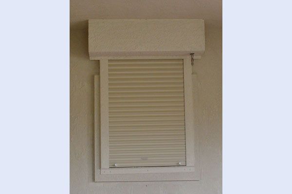 Beige window with closed, slatted, roll-up shade, framed in white, mounted on a beige wall.
