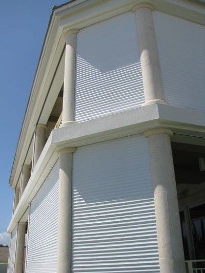 White roll-down shutters cover the windows of a two-story building with columns and a bright blue sky.