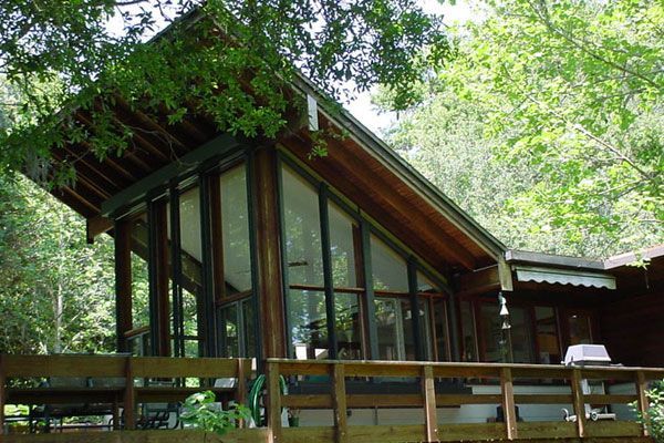 Wooden cabin with large angled windows, porch, and surrounded by trees.