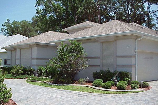 Tan house with a landscaped yard and paved driveway. Shutters cover the windows.