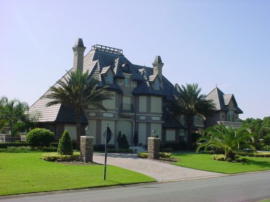 Large, ornate house with gray roof and stone facade, palm trees, and green lawn.