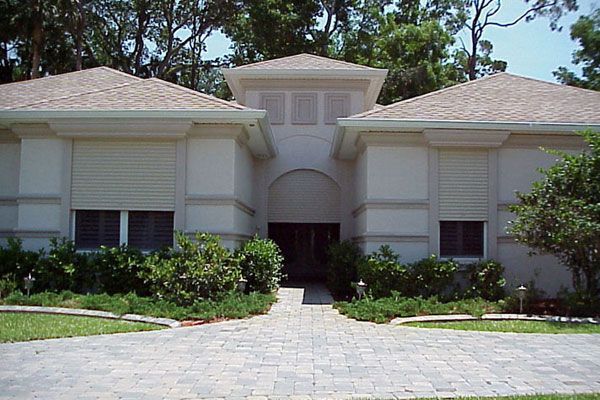 Beige house with roll-down shutters, brick walkway, and lush landscaping.