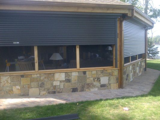 Exterior of a home with closed, gray roller shutters over windows. Stone wall and grassy lawn are visible.