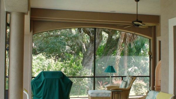 Screened porch with arched window overlooking greenery, with a grill, chair, and lamp inside.