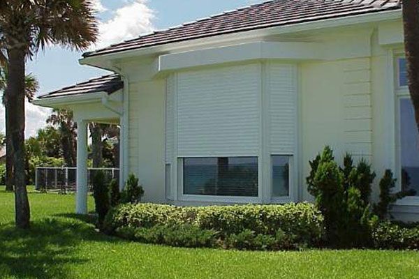 White house with closed security shutter over window, green grass, palm trees.
