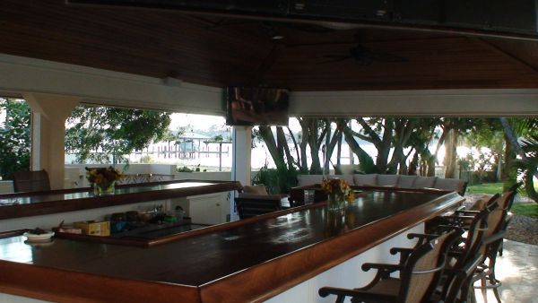 Outdoor bar with dark wood counter and stools, built-in appliances, and a mounted TV.
