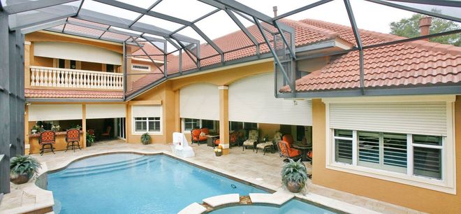A sunny, enclosed pool patio attached to a two-story home with neutral stucco walls and a reddish-brown tile roof.