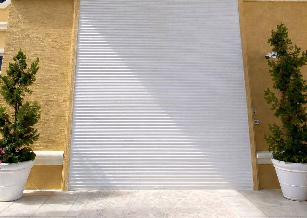 A white, horizontal-slatted metal roll-up garage door flanked by two potted cone-shaped evergreens against a yellow wall.
