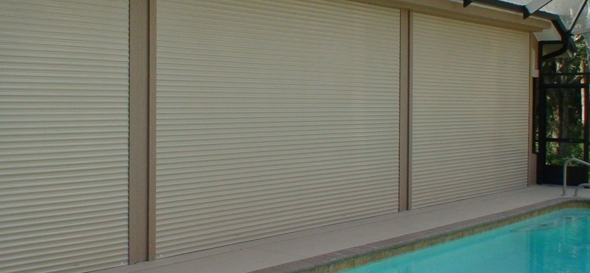 Beige rolling storm shutters fully closed over a large opening above a residential swimming pool deck.