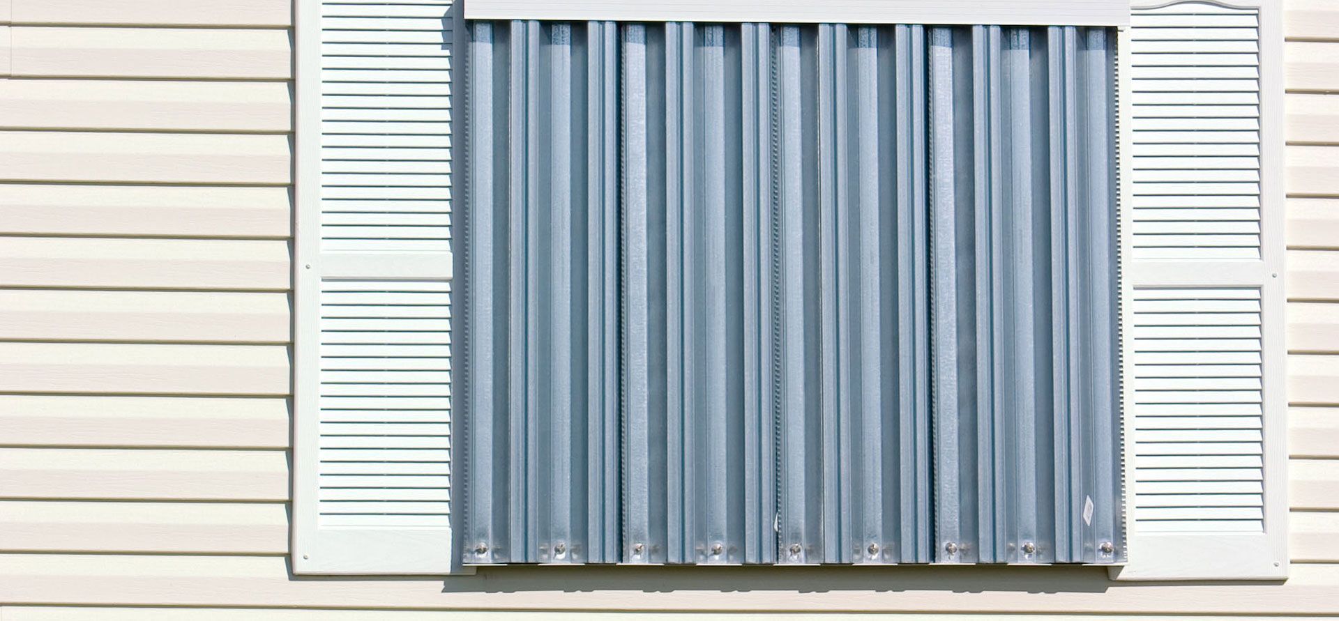 A window on a beige house covered with gray, vertical metal storm shutters and decorative white shutters on each side.