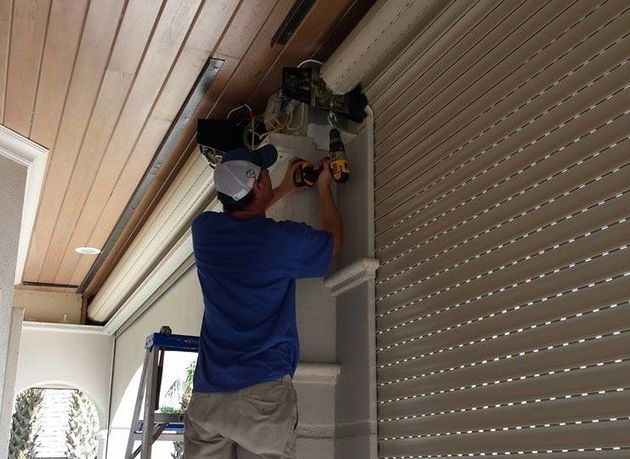 Man on a ladder installing a roll-up shutter with a drill, outdoors.