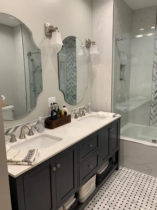 A bathroom with a dark blue double vanity, marble countertop, twin oval mirrors, wall sconces, and a glass-enclosed shower.