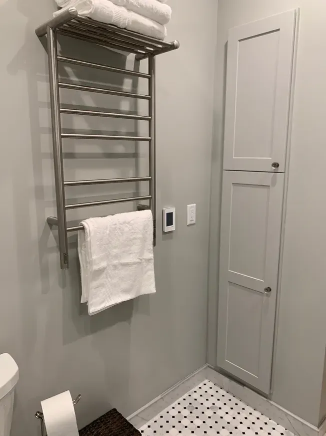 A stainless steel heated towel rack with stacked white towels on a grey bathroom wall next to a tall, narrow cabinet.