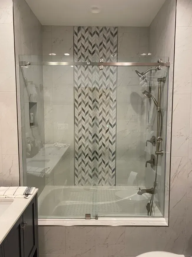 A glass sliding shower door over a white tub with a vertical decorative chevron tile accent wall and brushed nickel fixtures.
