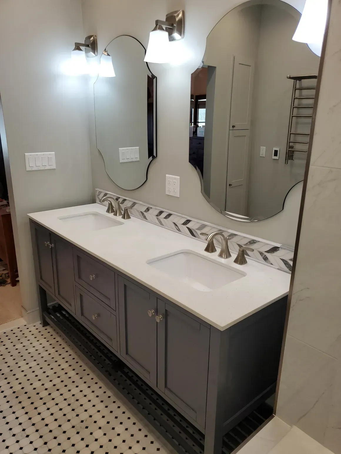 Double vanity with dark gray cabinets, white countertops, two decorative mirrors, and a herringbone backsplash.