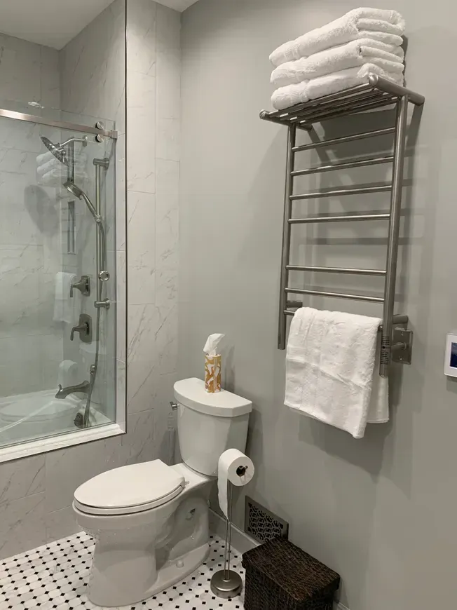 A bathroom with a toilet, a shower behind glass, and a metal towel rack on the wall holding stacked and hanging towels.