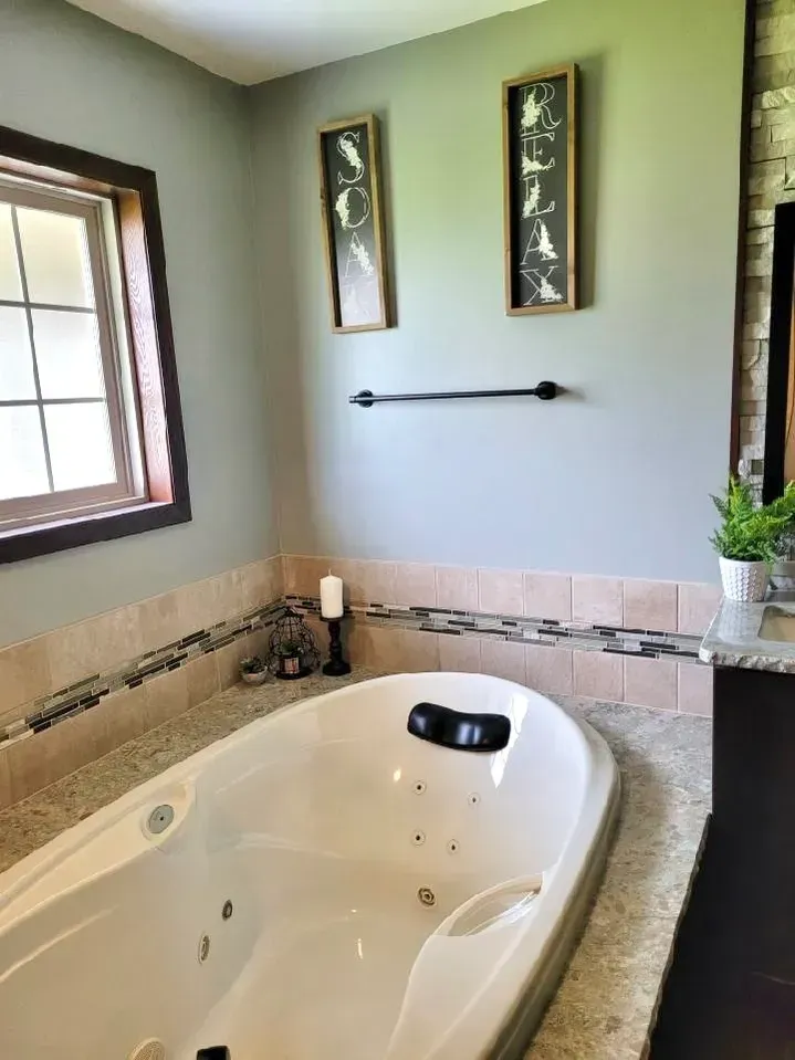 A soaking tub with a granite deck, set against a sage-green wall with two vertical art pieces and a black towel bar.