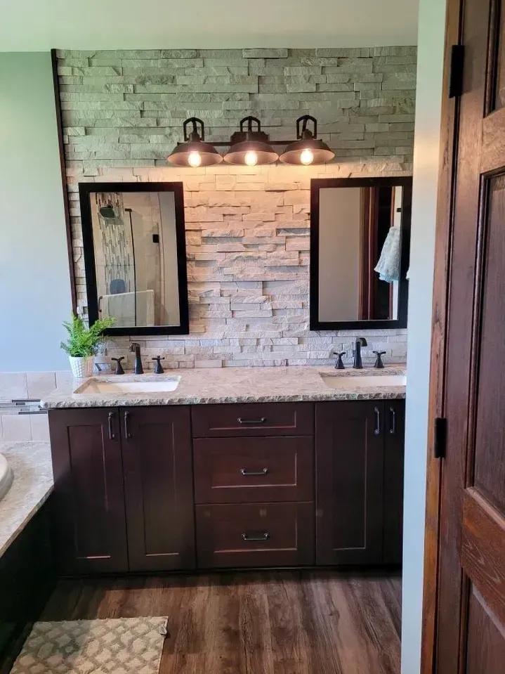 A bathroom vanity with a stacked stone backsplash, dark wood cabinets, a speckled countertop, and two framed mirrors.