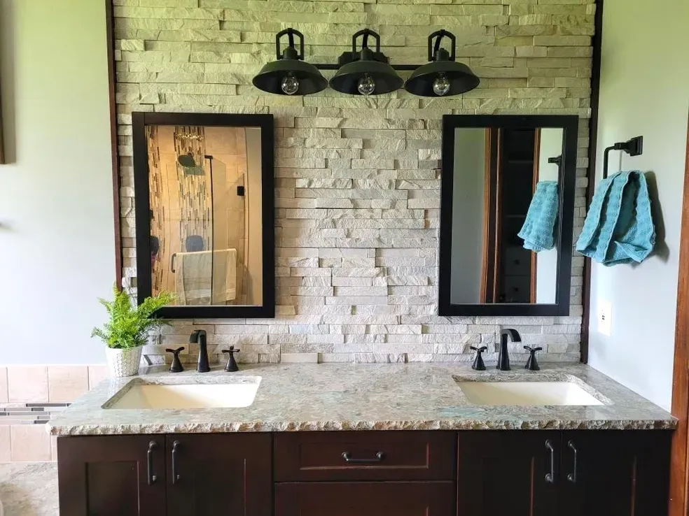 Double vanity with dark cabinets, stone tile backsplash, two mirrors, and overhead lighting in a bathroom.