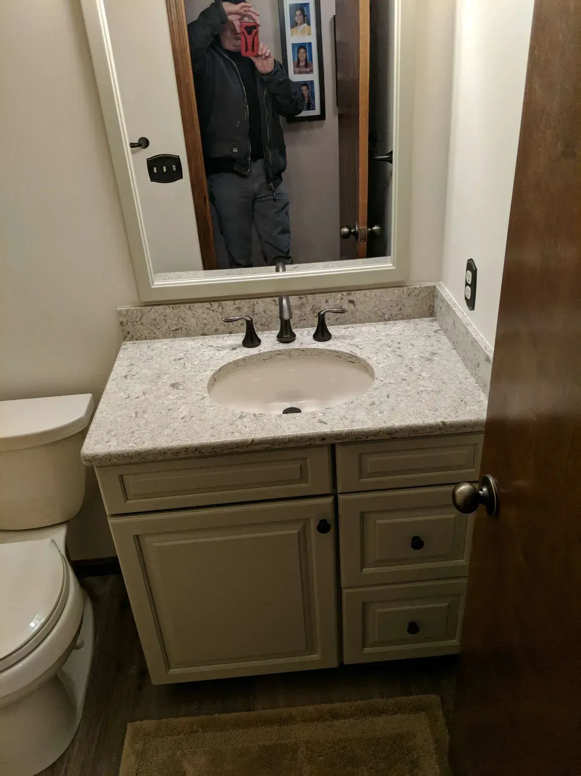 A bathroom vanity with a speckled white countertop, sink, and beige cabinet, with a mirror reflecting a person.