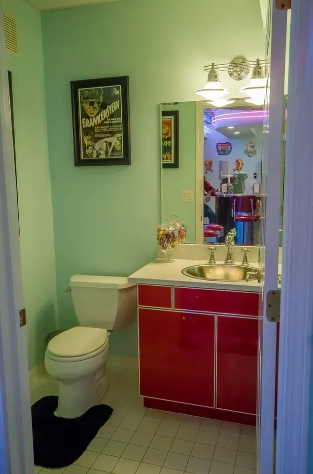 A mint green bathroom with a bright red vanity, a white toilet, a silver bowl sink, and a framed vintage poster.