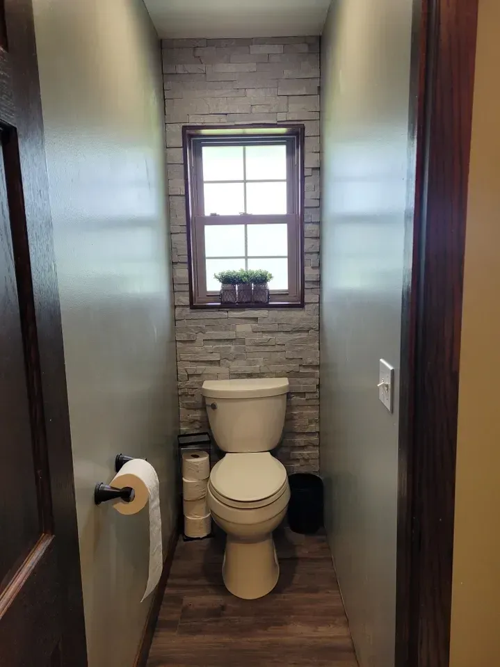A narrow, rustic-style water closet featuring wood flooring, neutral walls, and a stone-tiled accent wall behind a toilet.