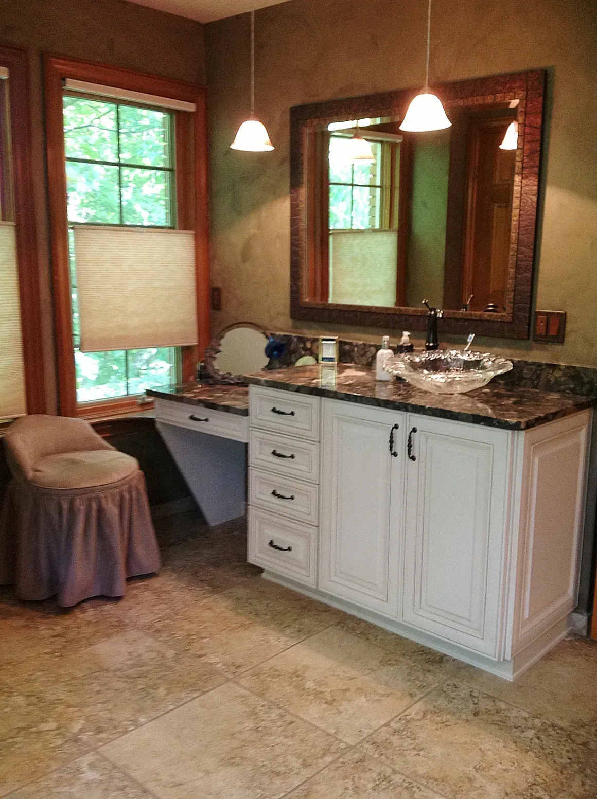 A bathroom vanity with a glass sink, white cabinets, a dark mirror, a makeup desk, and a stool near a window.
