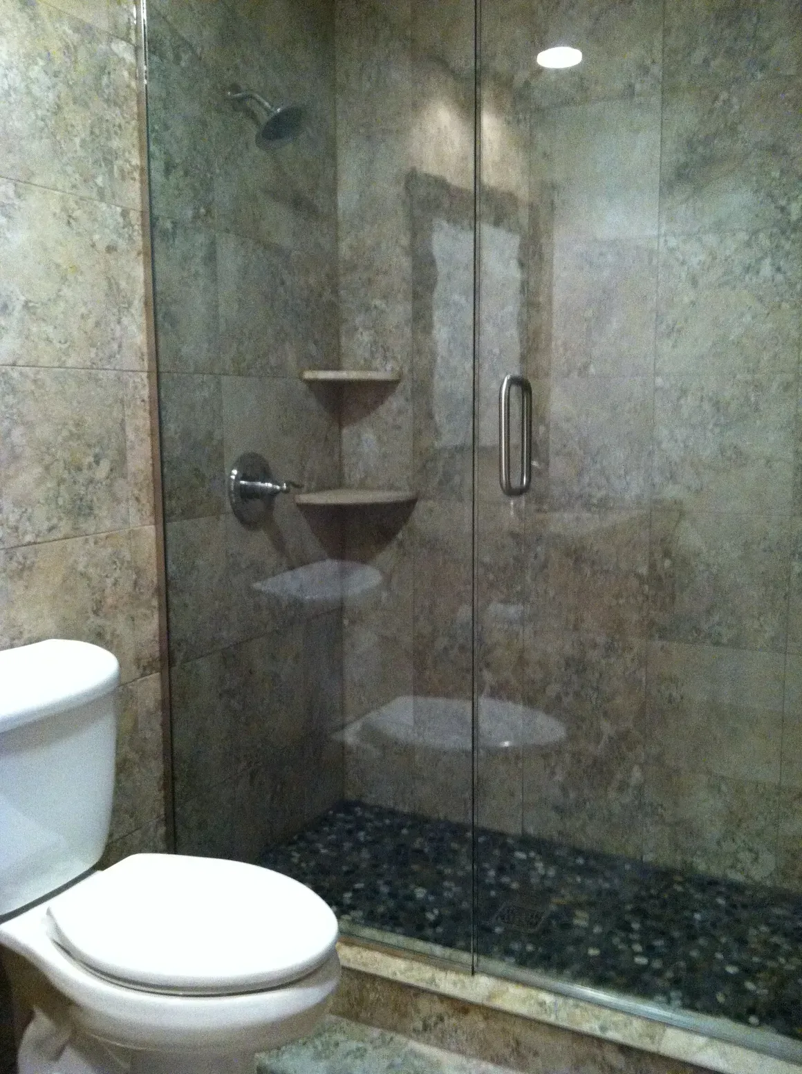 A white toilet sits beside a glass-enclosed shower with brown tiled walls and a dark pebble floor.