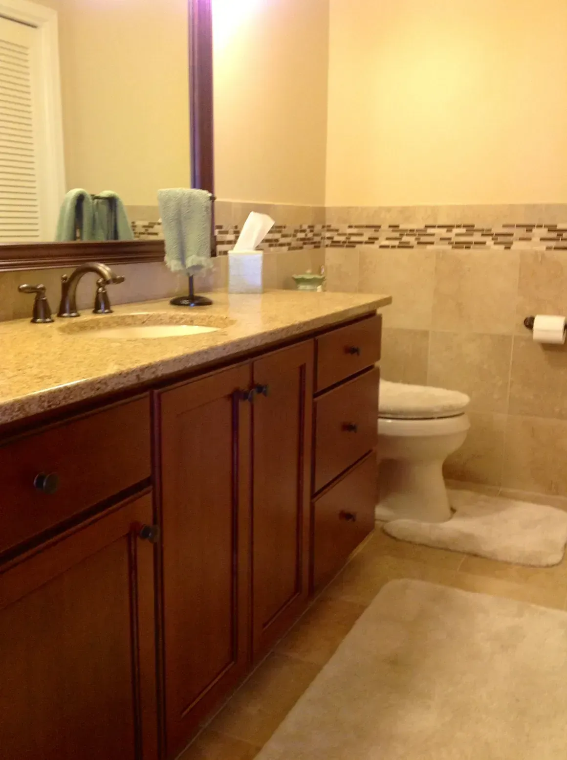 A bathroom with a cherry-wood vanity, beige stone countertop, tiled walls with an accent border, and a white toilet.