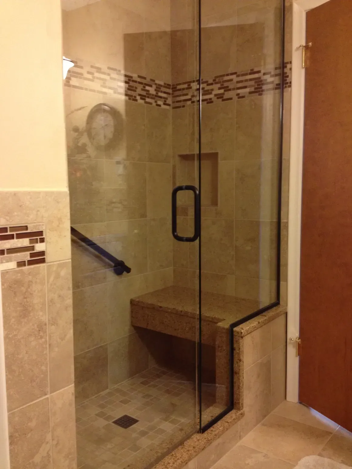 A walk-in shower with beige tile, a built-in bench, a dark metal grab bar, and a glass door with a black handle.