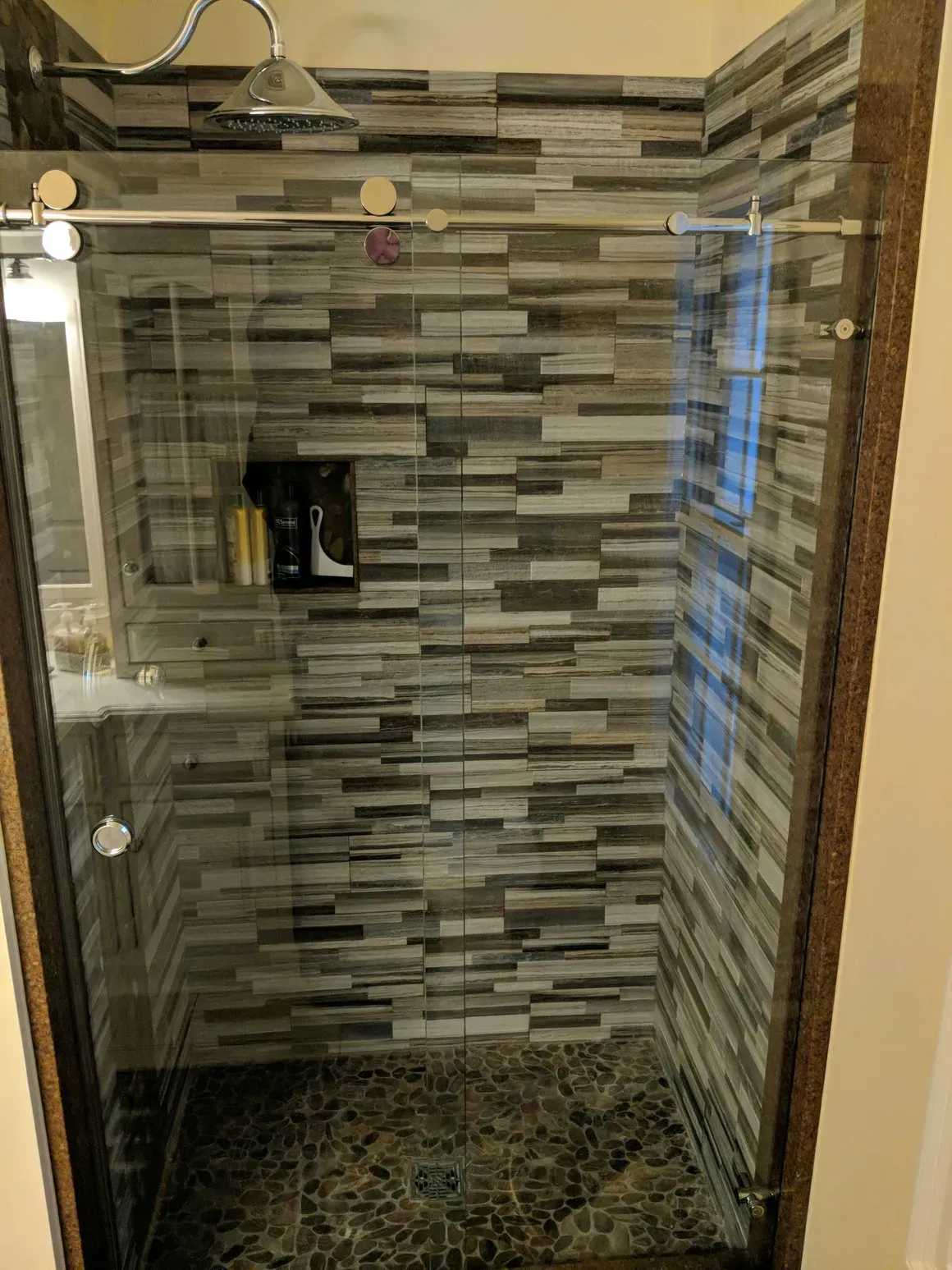 Walk-in shower with stone-look wall tiles, a glass sliding door, a rainfall showerhead, and a dark stone pebble floor.