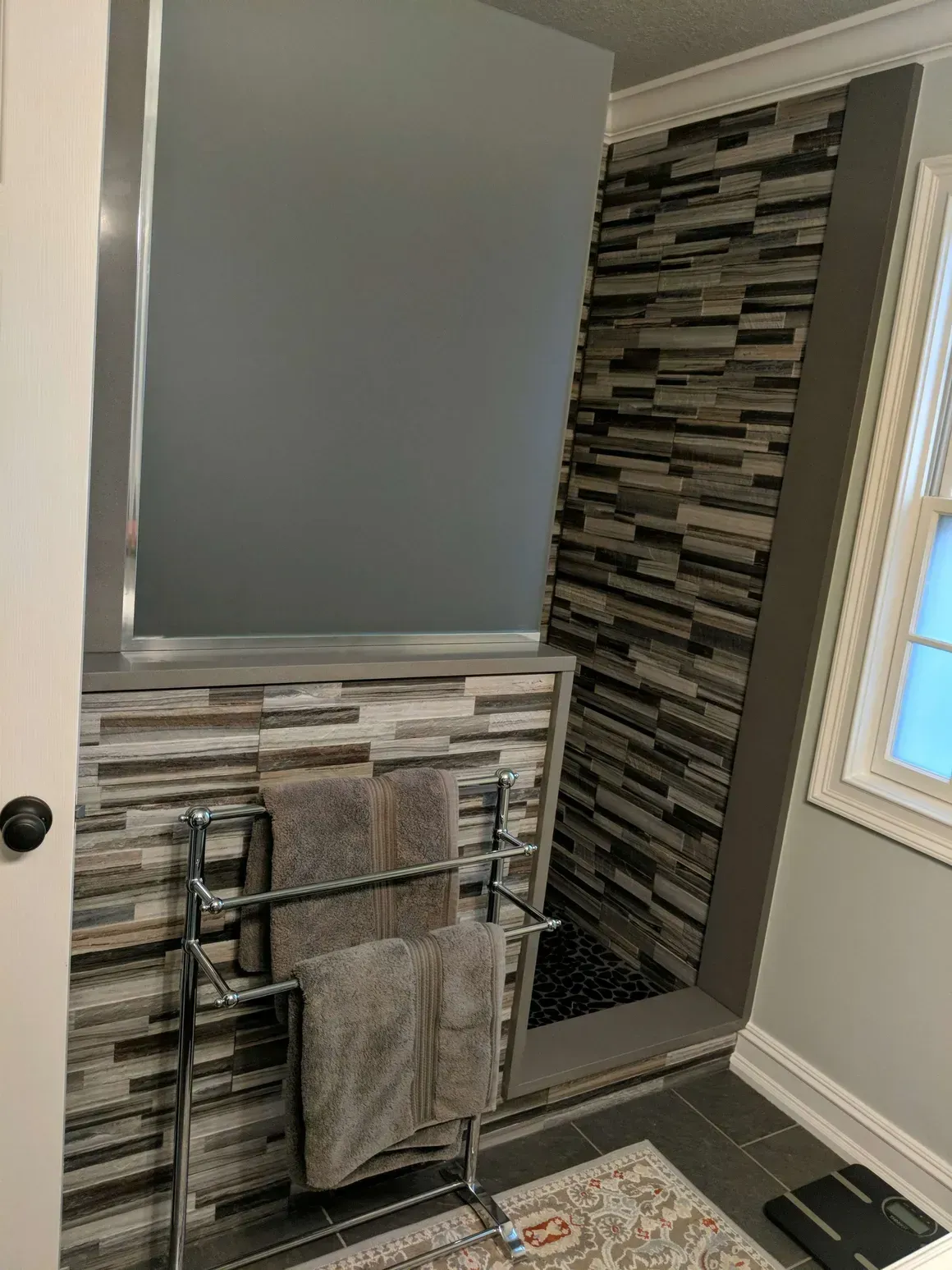A bathroom with a stone tile accent wall, a chrome towel rack with gray towels, and a window with white trim.