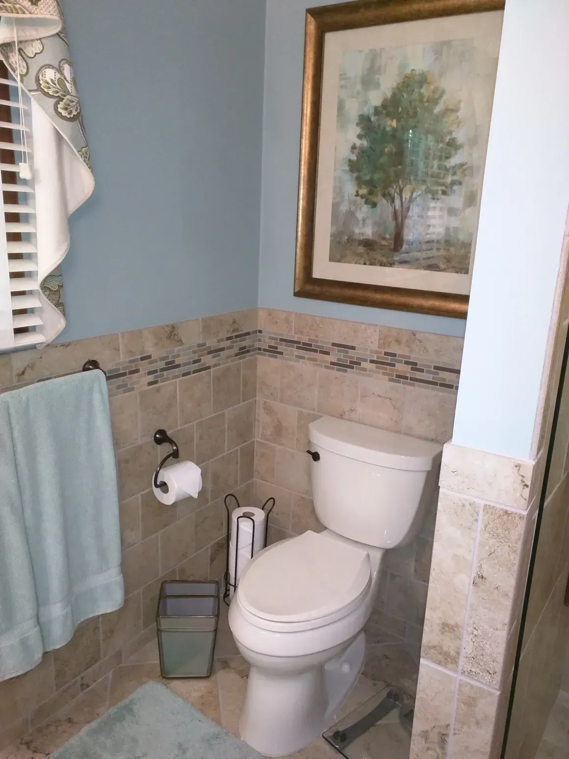 A powder room featuring a toilet, a neutral tiled wall, a light blue hand towel, and a framed tree painting.