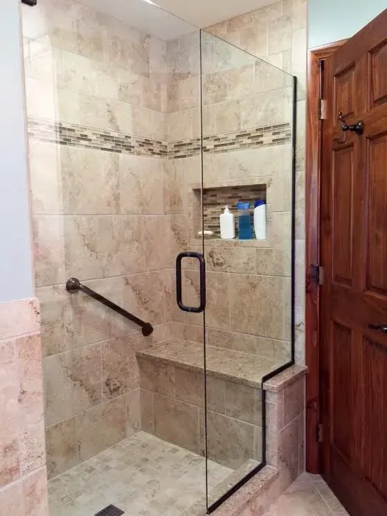 A modern tiled walk-in shower with a glass door, built-in seat, a recessed shelf, and an installed safety grab bar.