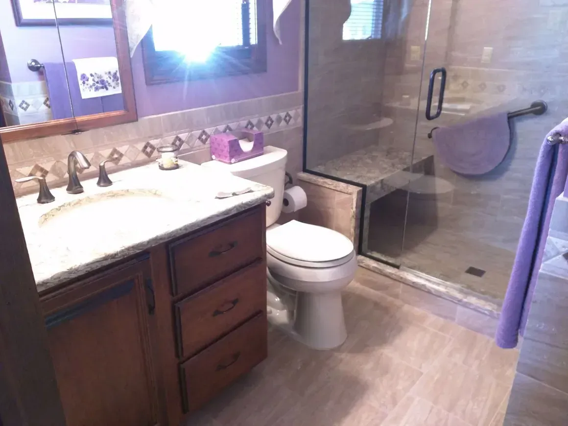 A bathroom with a wooden vanity, a light-colored countertop, a white toilet, and a glass-enclosed shower with purple towels.