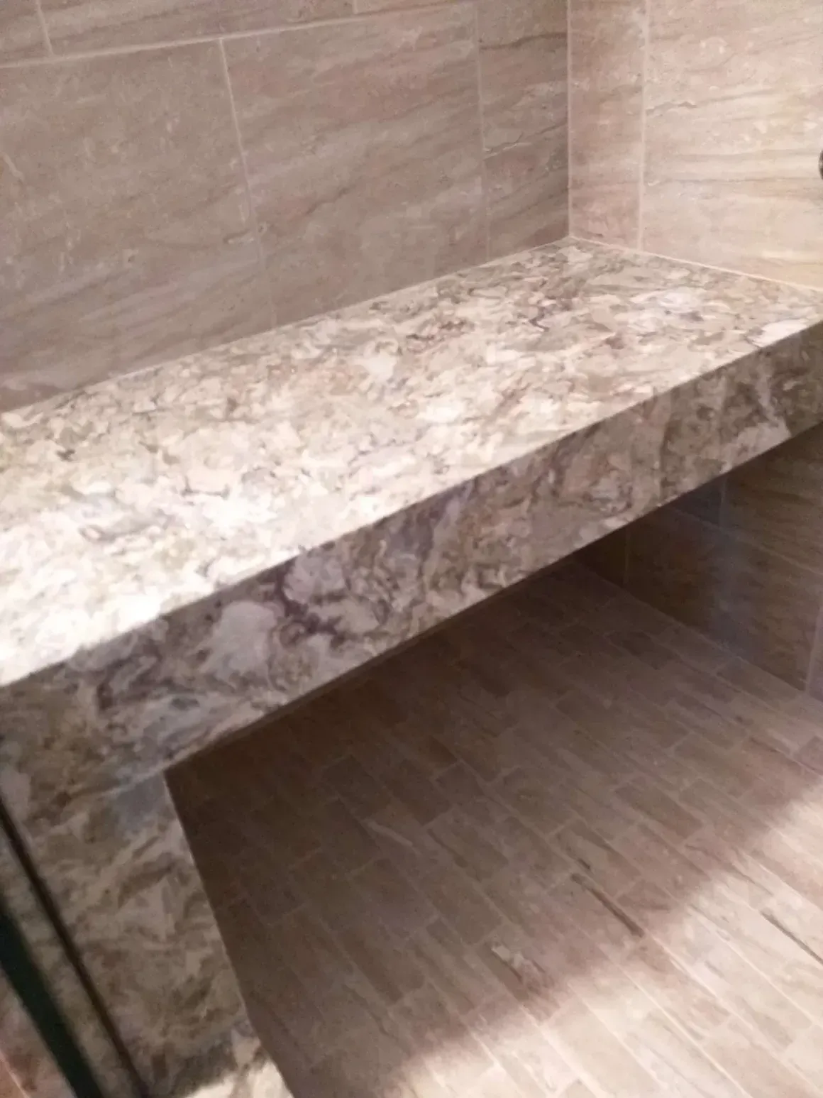 A marble shower bench with a thick slab top and base, set against light-colored, vertically stacked tiled walls.