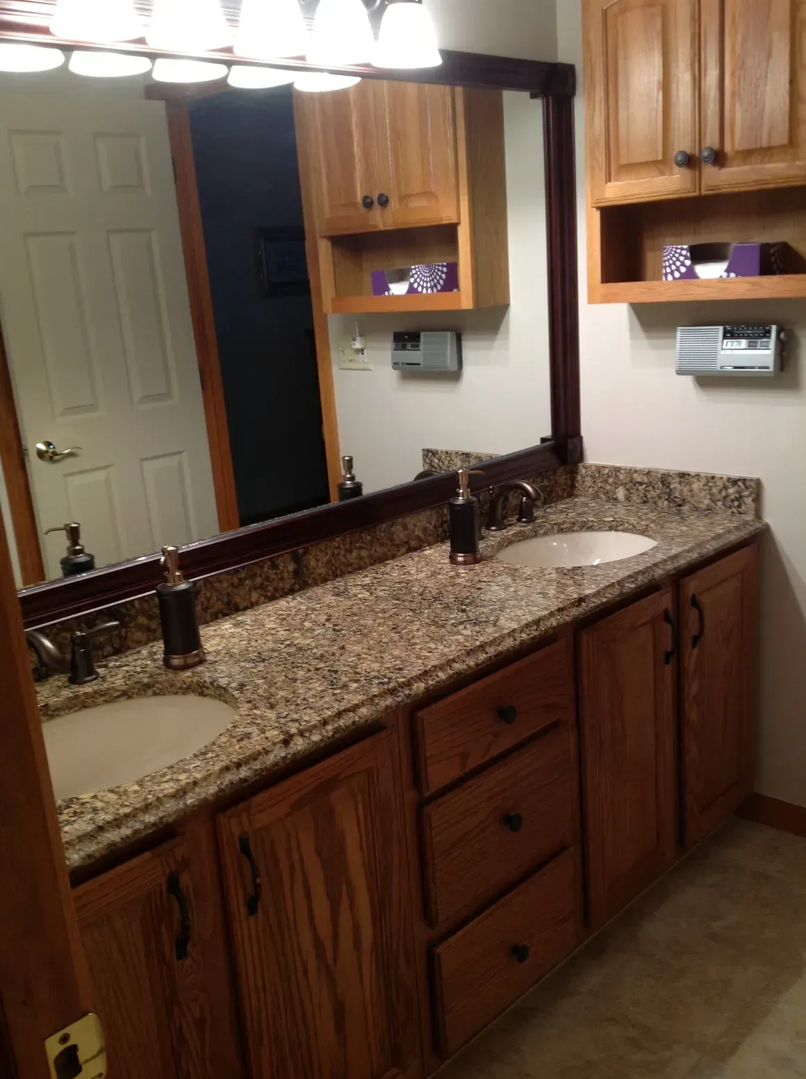A bathroom vanity with oak cabinets, a granite countertop, two sinks, and a large mirror with a dark wood frame.