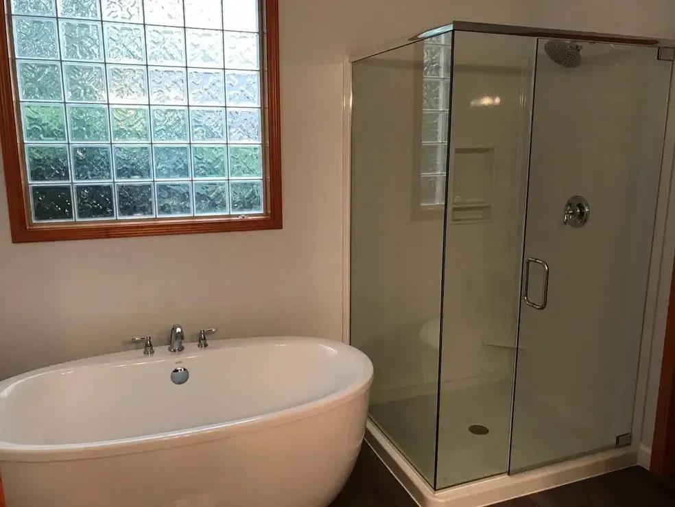 A modern bathroom featuring a white freestanding soaking tub, a glass-enclosed walk-in shower, and a glass block window.