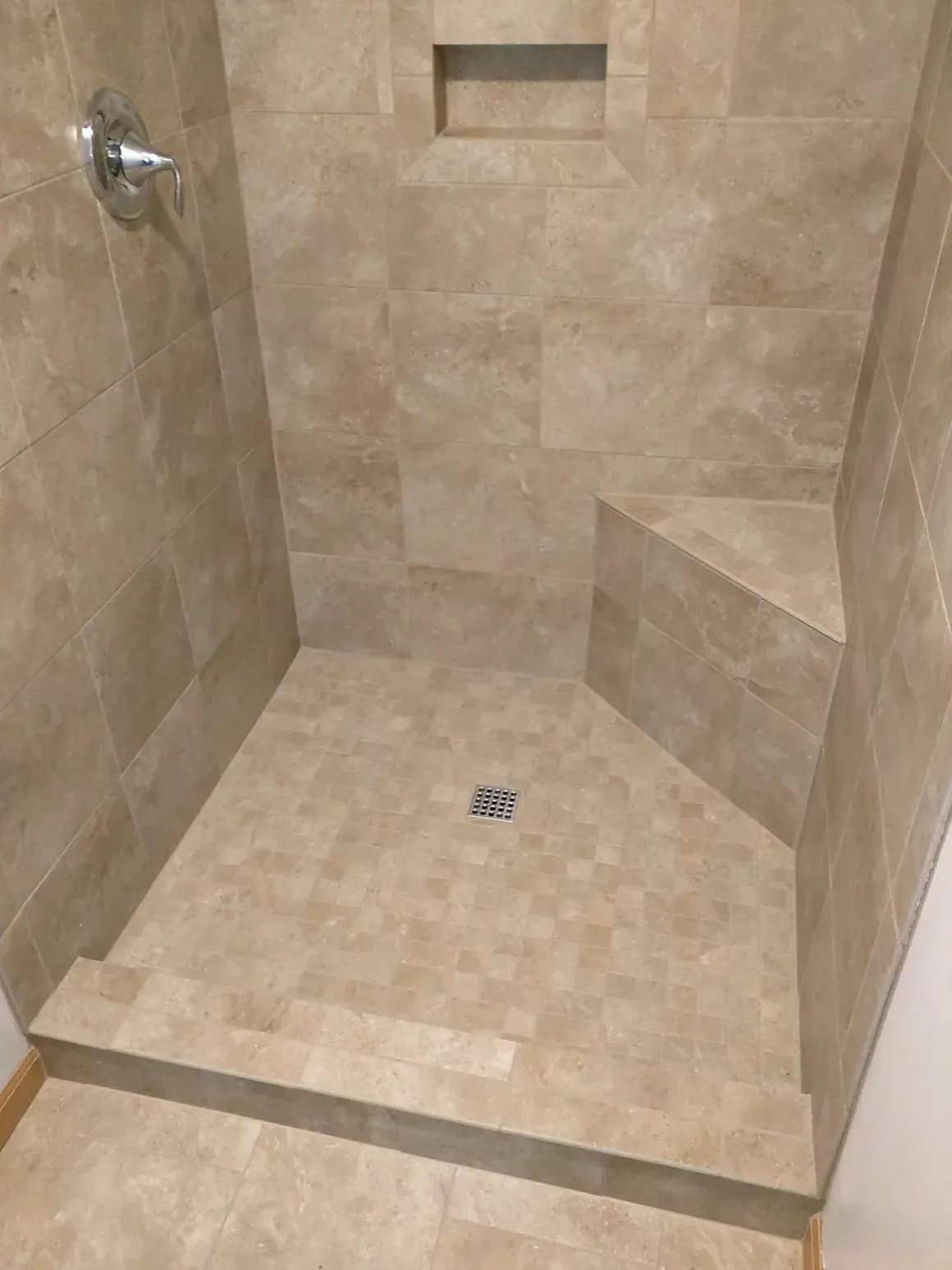 A beige tiled walk-in shower with a built-in corner seat, shower niche, and a low step-in threshold.